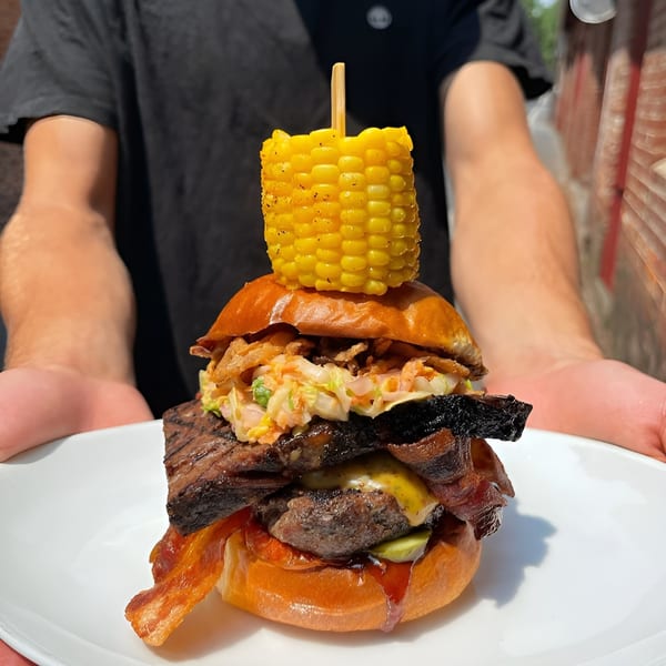 A person holds a large Maxwell's burger topped with corn on the cob, bacon, and coleslaw.