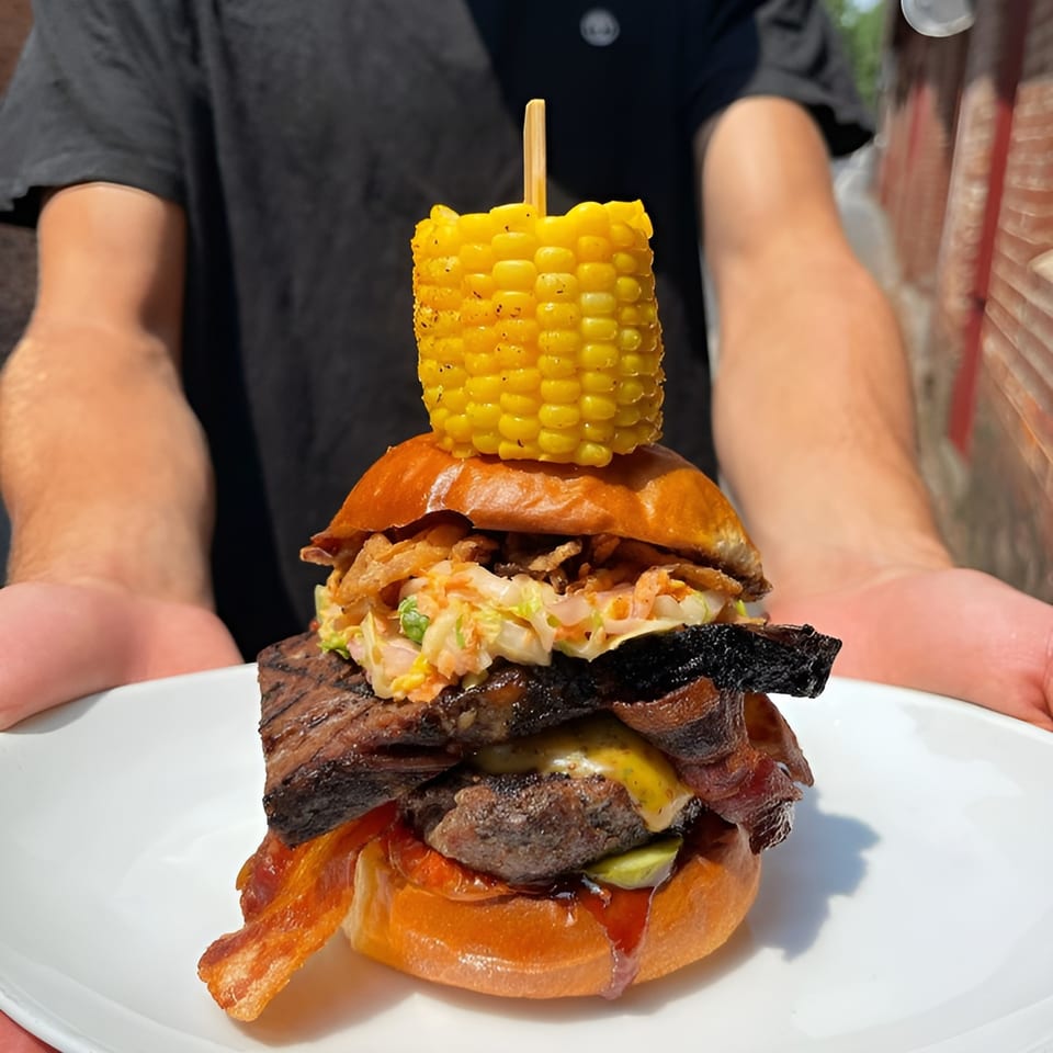 A person holds a large Maxwell's burger topped with corn on the cob, bacon, and coleslaw.