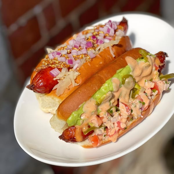 Two gourmet hot dogs on a white plate, one with relish and mustard, the other with avocado and salsa.