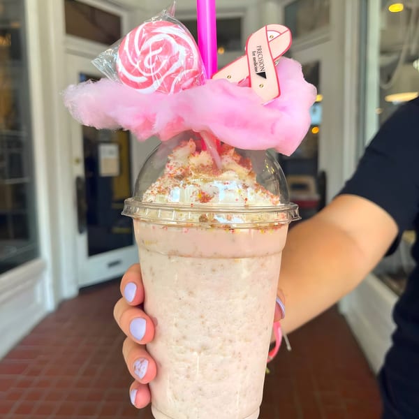 Hand holding a Maxwell's Burgers & Shakes milkshake topped with whipped cream, sprinkles, cotton candy, and a lollipop.