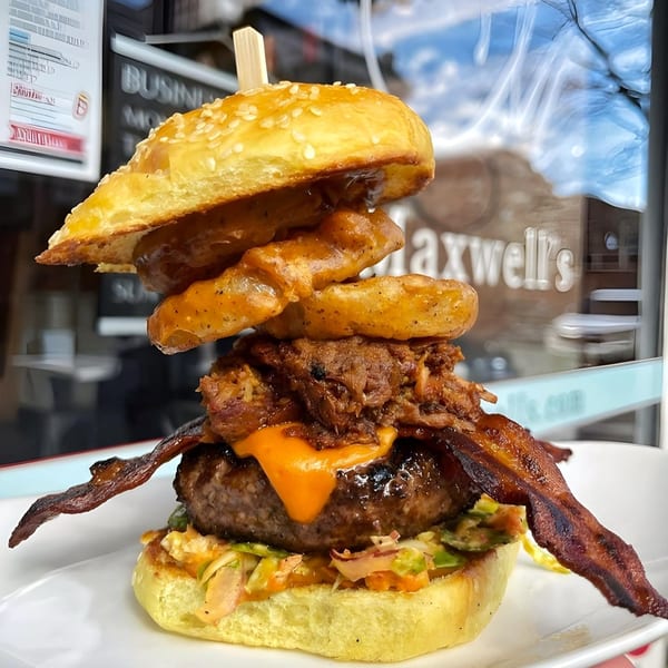 A tall Maxwell's burger with pulled pork, onion rings, bacon, and cheese sauce on a sesame bun.