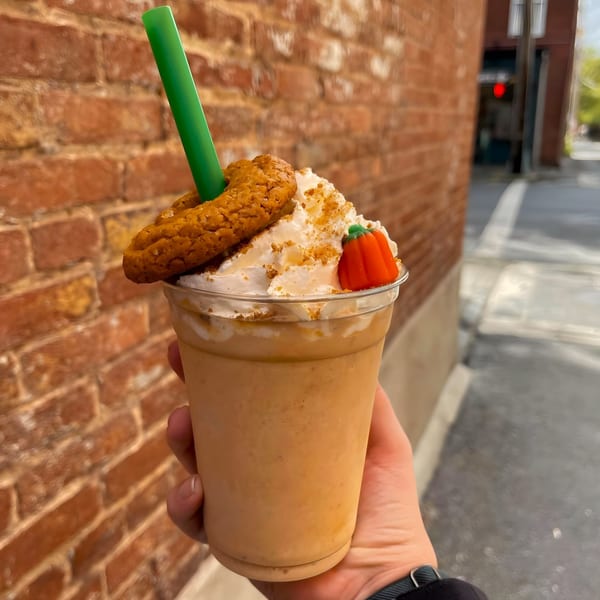Hand holding a Maxwell's pumpkin shake with whipped cream, cookie, and candy pumpkin against a brick wall.