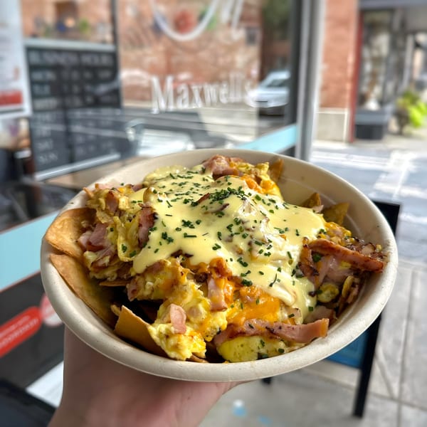 A hand holds a bowl of breakfast nachos with scrambled eggs, ham, cheese sauce, and chives.